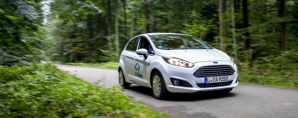 Ein Auto fährt auf einer Straße, rechts und links Wald, auf dem Auto ist ein teilAuto-Logo