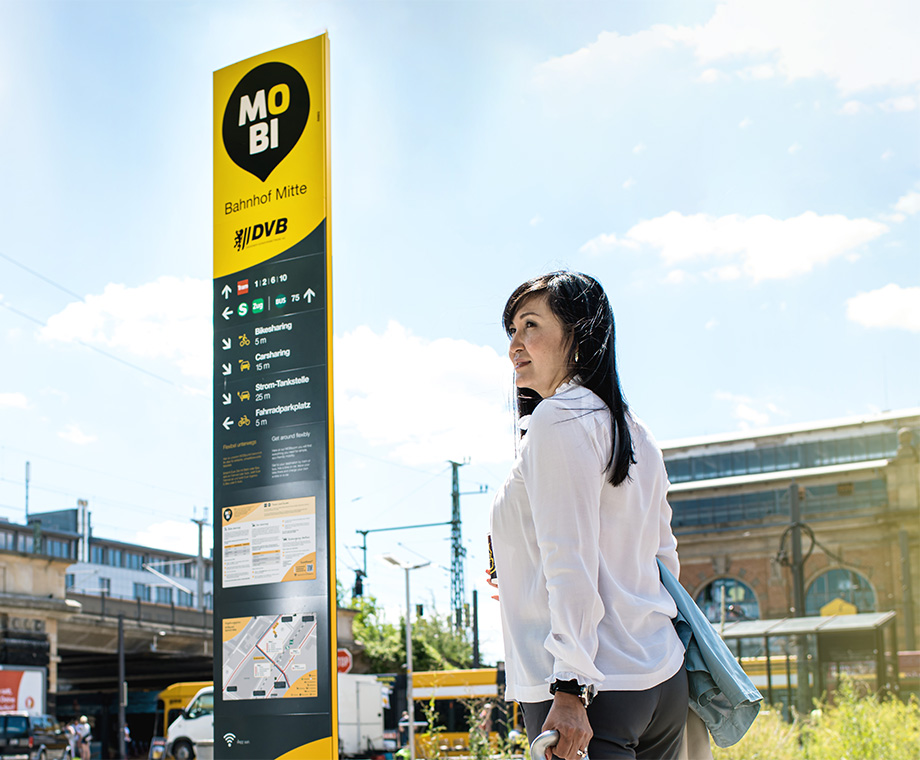 Eine Frau steht mit einem Koffer an einem Mobipunkt vor einem Bahnhof